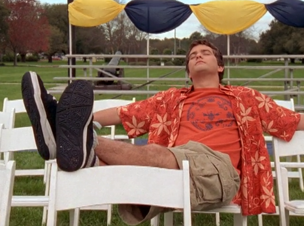 Pacey reclines in a graduation chair in an empty row, wearing a bright orange Hawaiian shirt and a very satisfied look on his face