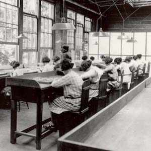 A group of young women sit at desks in a factory