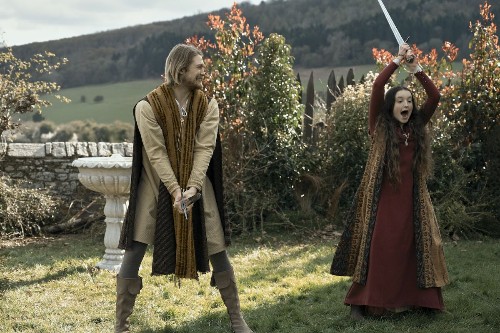 A handsome white man with blond hair and a beard (Joe Alwyn) grinning at a young white girl (Bella Ramsey) who is triumphantly brandishing a sword