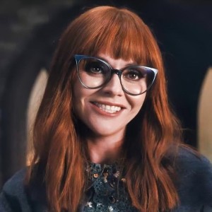 A white woman with long auburn hair and bangs, wearing blue framed cats eye glasses and smiling awkwardly