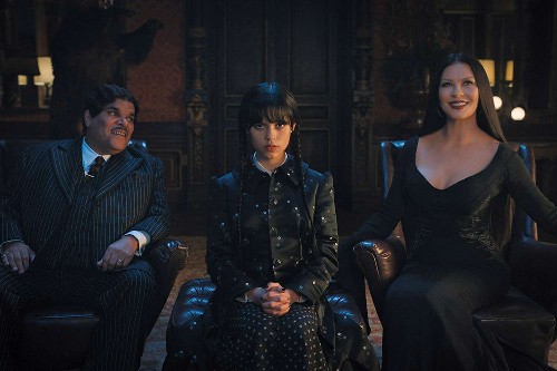 A grinning Puerto Rican man with a thin mustache and shiny black hair wearing a pinstripe suit and a glamorous woman with long dark hair and a black dress, with Wednesday (scowling girl with dark hair in braids) sitting between them