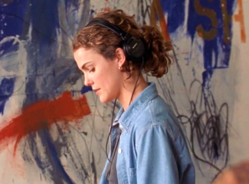 Felicity stands in front of a large abstract painting she's working on, looking down with headphones on