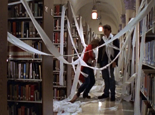 Felicity and Noel stand, laughing, in a library covered floor to ceiling with strands of toilet paper