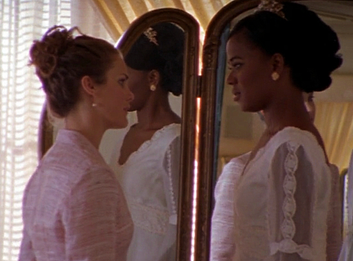 Felicity and Elena stand in front of a full-length mirror looking at each other with intent expressions, Felicity in a pink top and Elena looking breathtaking in a wedding dress