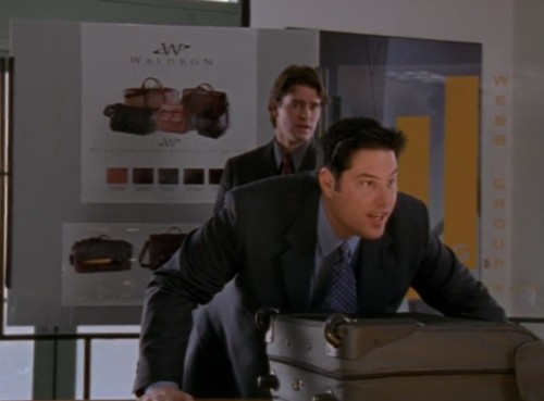 Sean, in a suit, is presenting a suitcase while Noel, also in a stand, stands behind him looking concerned