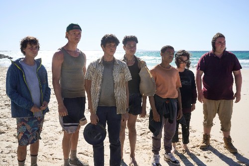 A row of teenage boys standing together, looking bedraggled, on a beach.