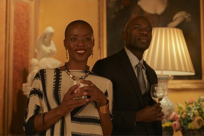 A gorgeous Black woman with closely shaved hair, holding a glass, stands next to a handsome Black man in a suit
