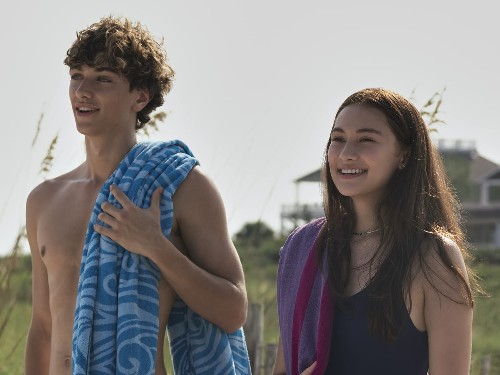 A fit white blonde teenage boy, shirtless with a towel over his shoulder, stares at the ocean next to a biracial (half Asian, half white) teenage girl in a swimsuit 