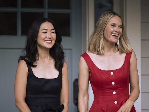 A middle aged Asian woman with long hair in a black sundress stands on a porch, smiling, next to a white blonde woman, also smiling, in a red sundress