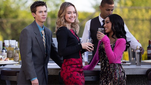 Three teenagers stand together at an outdoor bar: a slender white guy with brown hair, a pretty blonde white girl, and a pretty Black girl smiling at the other girl