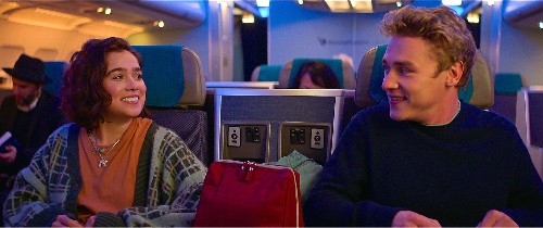 A pretty white girl with brown curly hair, sitting in an airplane seat, smiles at the white, blonde, cute boy sitting next to her