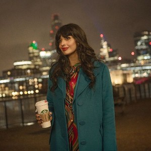 A gorgeous, brown-skinned woman with long dark hair and bangs, wearing a blue coat and holding a coffee with a city skyline in the background