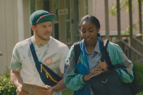 A young white man wearing a flipped up cap and fanny pack talking to a young Black woman holding a bag