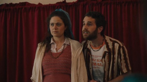 Photo of Molly Gordon, a white young woman with long brown hair, standing in front of a red curtain next to Ben Platt, a white young man with brown hair and a beard