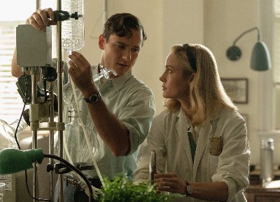 A handsome young white man (Lewis Pullman) in a lab coat looking at a pretty white blonde woman (Brie Larson) in a lab coat, sitting in a chemistry lab