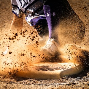 A person in purple baseball gear slides into a base in a cloud of sand