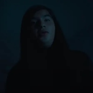 A youn gman with long black hair looms in the dark