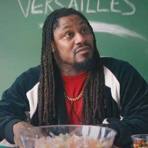 Marshawn Lynch, a broad shouldered Black man with long braids sitting at a desk