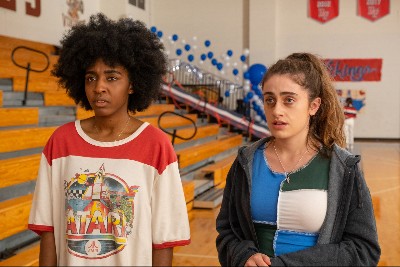 Ayo Edebiri, a young Black woman with an Afro, and Rachel Sennott, a young white woman with a frizzy ponytail and a black eye, stand in a high school gym looking worried