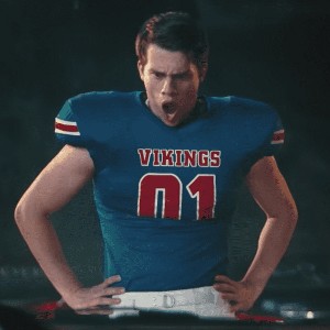 Nicholas Galitzine, an attractive young white man wearing a blue and red football jersey and making a shocked expression