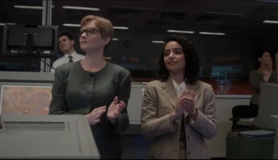 A tall white woman and a shorter Latina woman stand together clapping in NASA Mission Control.