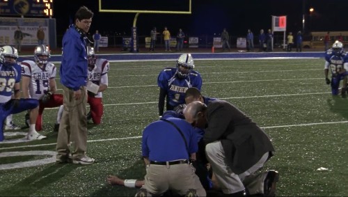 A screenshot from the pilot, with Coach Taylor on the football field looking down as officials and medics kneel around a fallen player