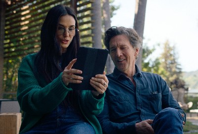 Photo of Demi Moore, a striking white woman in her early 60s with glasses and long dark hair, looking at an iPad with Andrew McCarthy, a white man with swoopy gray hair, also in his early 60s