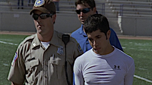 A white man in a Sheriff's uniform escorts a handcuffed Hispanic teenager with a white male football coach behind them.