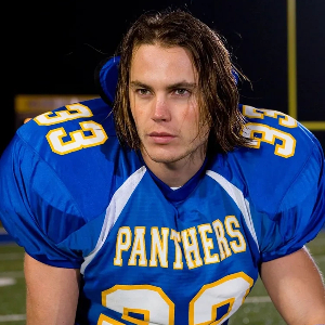 Tim Riggins, a handsome white guy with messy, chin-length brown hair, wearing a blue Panthers football jersey