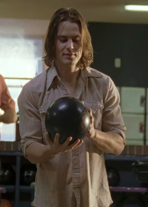 Tim Riggins, a handsome white boy with shaggy brown hair, stands in a bowling alley, holding his bowling ball, ready to roll it, lookign down in concentration with a smile on his face