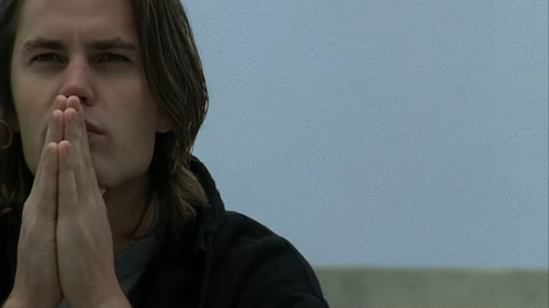 Tim Riggins, a white boy with brown hair, poses pensively with his hands held together in front of his face.