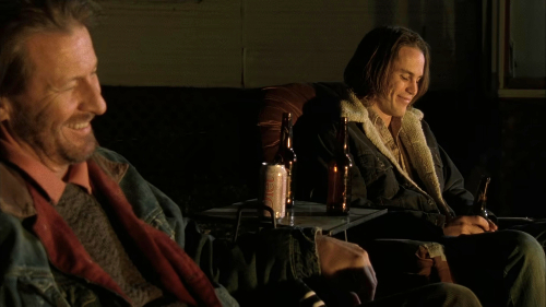 Tim Riggins and his dad, a white teenage boy and his father, sit around a fire with beer bottles on the table between them, smiling