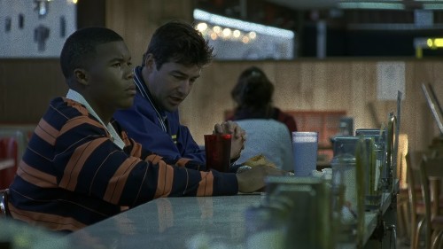 Smash, a young Black man, sits at a diner counter next to Coach Taylor, a white man in a coaching jacket