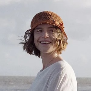 Jessie Buckley as Rose Gooding in Wicked Little Letters wears a white dress and a brown hat on the beach