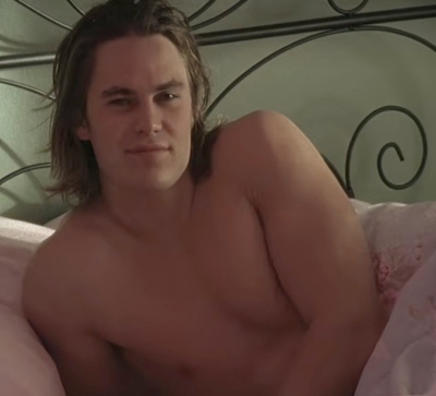 Tim Riggins, a white teenage boy with long shaggy brown hair and a smile reclines shirtless on a bed