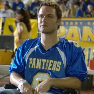 Jason Street, a handsome white guy with short brown hair, sits in a wheelchair wearing a blue Panthers football jersey