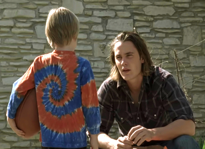 Taylor Kitsch as Tim Riggins, squatted down to talk to a kid holding a football with his back turned to the camera