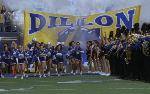 A group of cheerleaders stand in front of a Dillon banner as football players break through it on the field