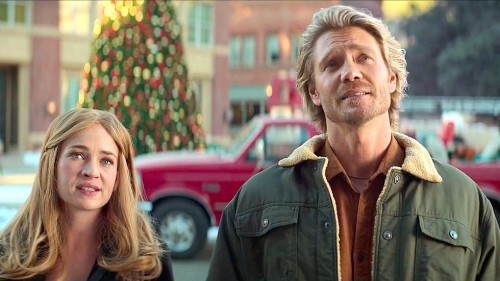 A pretty white blonde woman stands next to a handsome white man with a Christmas tree in the background