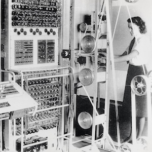 A black and white photo of a woman from the 1940s standing in front of a giant computer.