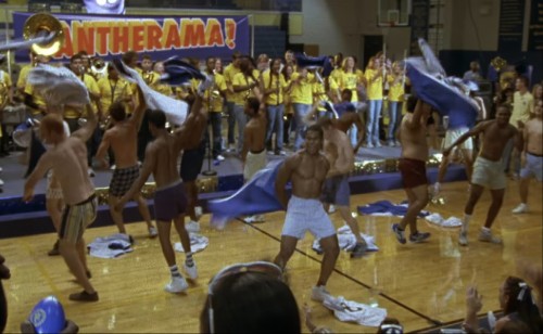 A pep rally in a high school gym with football players shirtless, waving their pants around