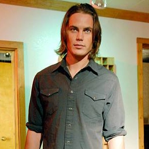 a teenage white boy with shoulder length hair, wearing a button-up shirt, stands in a hallway