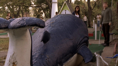 A putt-putt course with a giant whale sculpture in the foreground. In the background, Minka Kelly as Lyla Garrity laughs with Matt Czuchry as Chris.