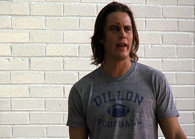 Taylor Kitsch as Tim Riggins, standing against a white brick wall, wearing a grey Dillon Football t-shirt. His brown hair is long and his face is kind of beat up