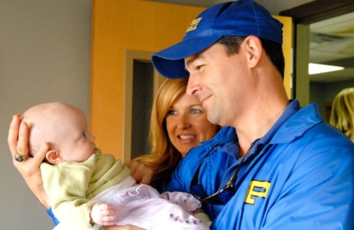 a white man in a blue jacket and cap holds a small baby in his hands while a blonde white woman smiles over his shoulder