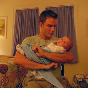 Scott Porter as Jason Street on Friday Night Lights. He's a handsome young white man with short brown hair, sitting in a wheelchair in a bedroom, holding a small baby and looking at him with love.