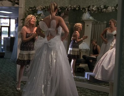 Mindy trying on a white wedding ballgown with small wings on the back while her mom looks on proudly