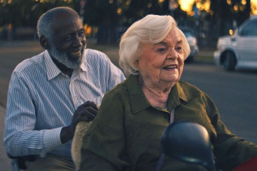 An elderly black man sits behind an elderly white woman on a scooter, both are laughing