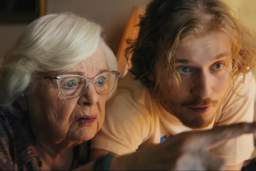 An elderly white woman with white hair and glasses and a young man with shaggy blonde hair look at a screen together