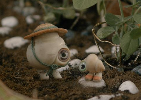 A large shell wearing a hat with one googly eye stands next to a smaller shell with one googly eye in a garden